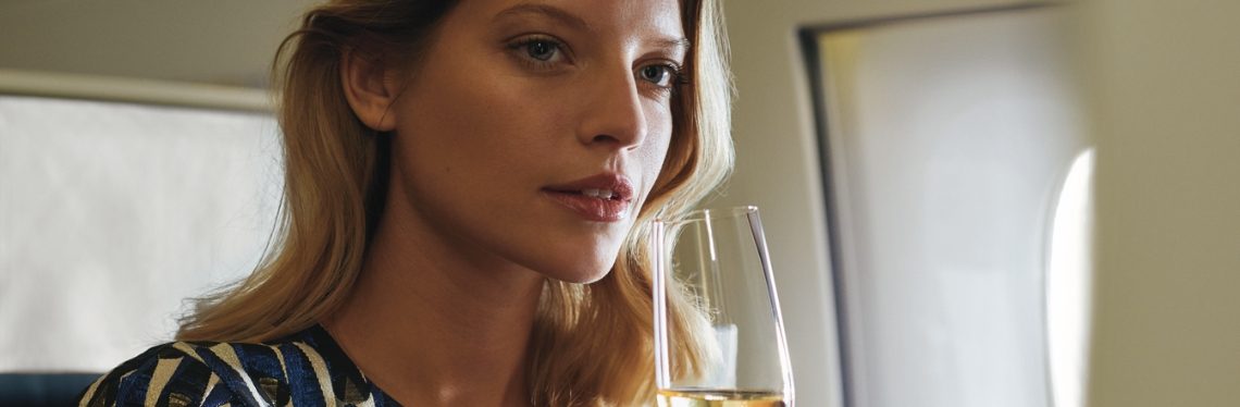 A woman on an airplane is sitting by the window in First Class, holding a glass of champagne.