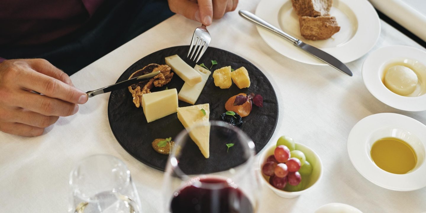 Eine Person in der ersten Klasse eines Flugzeugs geniesst eine Käseplatte mit verschiedenen Beilagen. Auf dem Tisch stehen zudem ein Glas Rotwein, Brot, Olivenöl, Walnüsse und weitere kleine Schälchen mit Speisen.